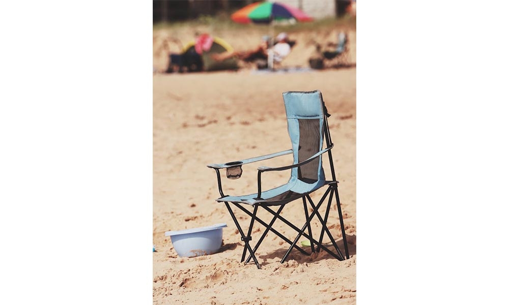 Blue folding chair on beach with a bucket beside it, beach umbrella in background